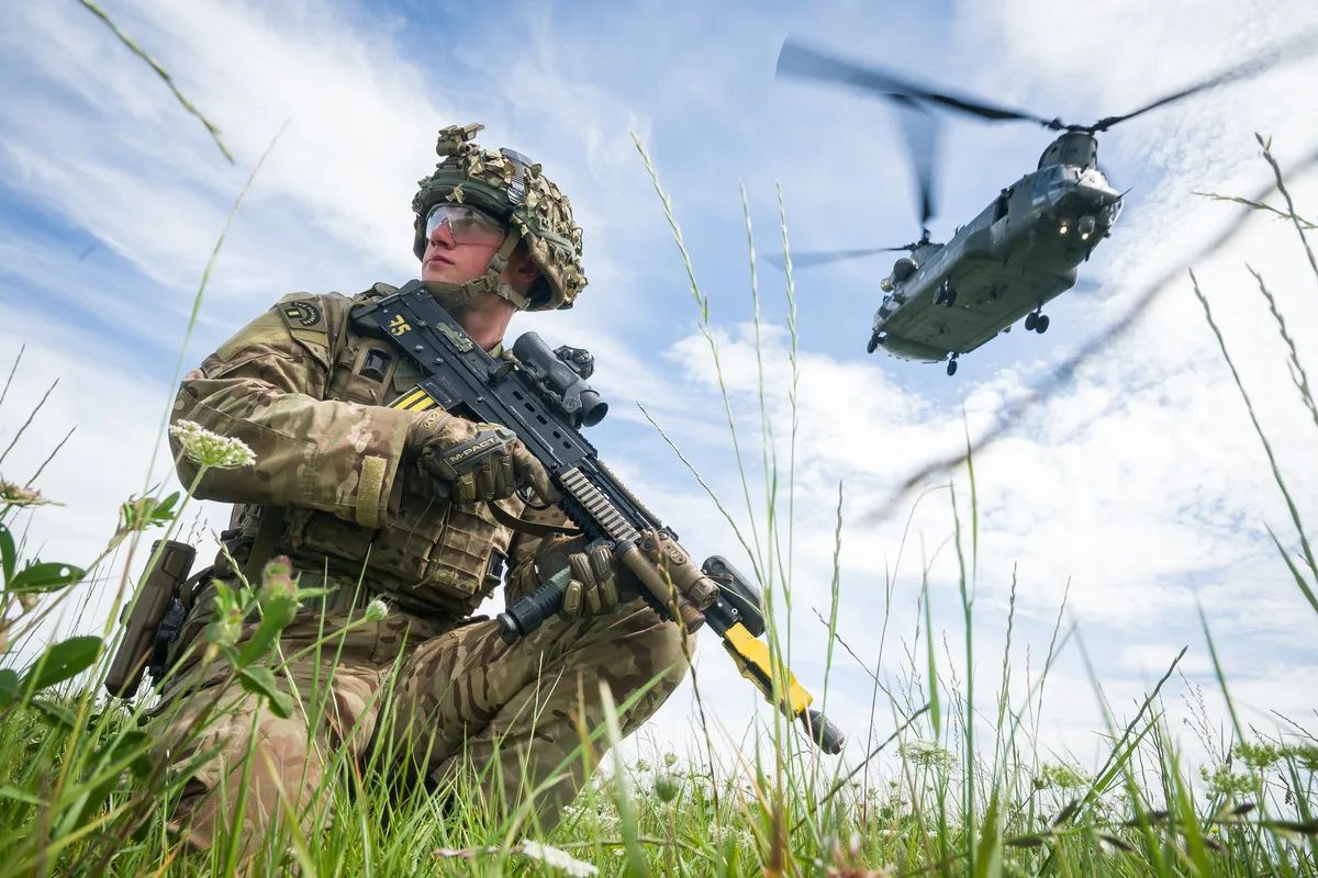 A soldier kneeling in long grass as a Chinook helicopter flies past
