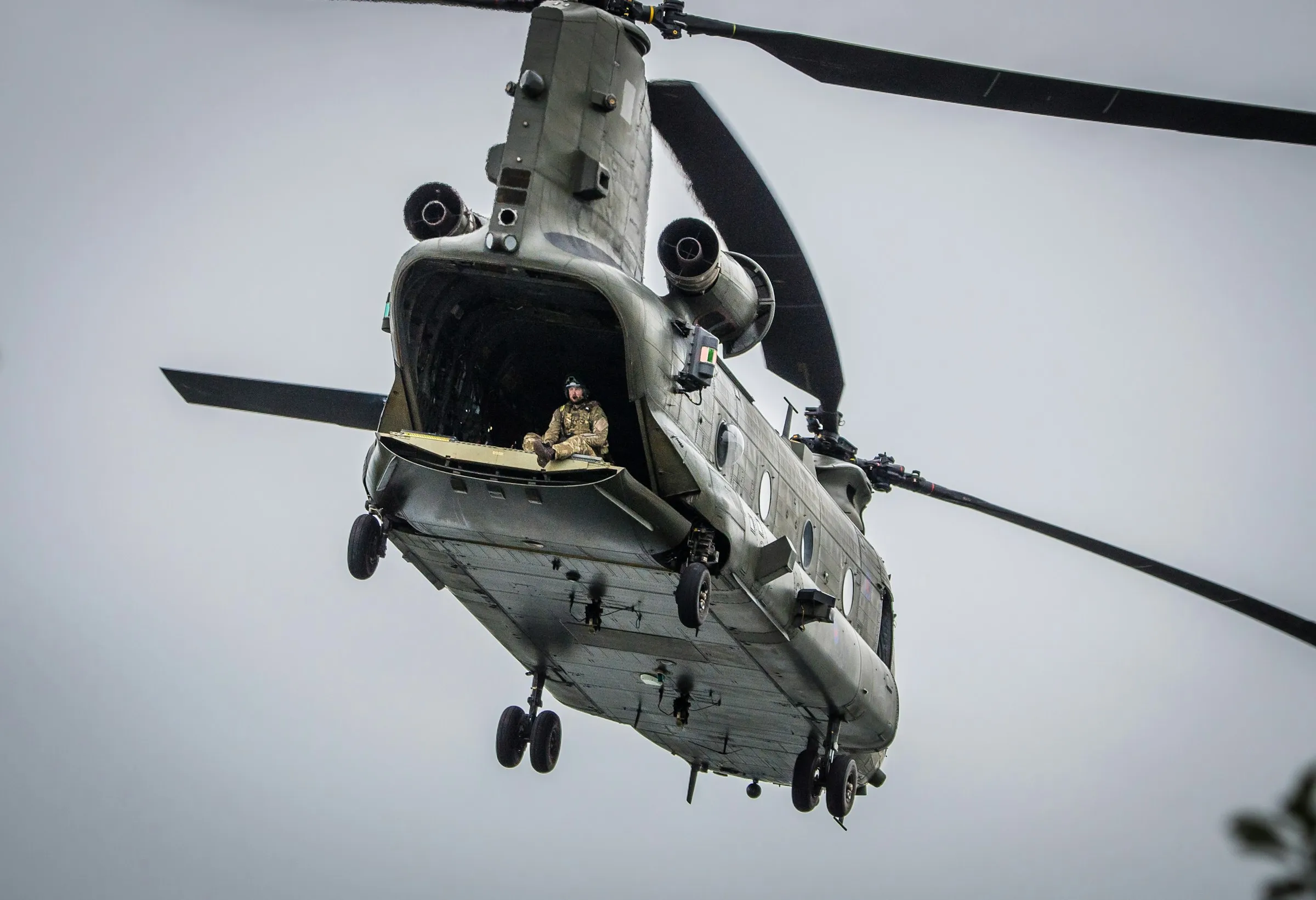 An RAF Chinook helicopter in flight with a crew member seated on the open rear ramp